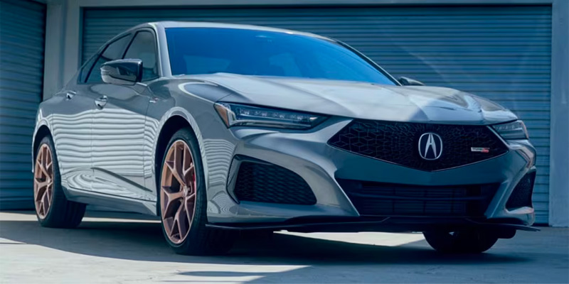 A sleek gray 2025 Acura TLX sedan with a sporty design and bronze wheels, parked in front of a garage.