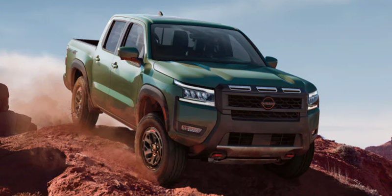 A green 2025 Nissan Frontier pickup truck climbing a rocky terrain under a blue sky.