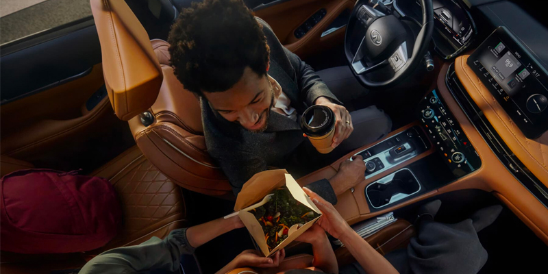 Two people in a stylish 2025 INFINITI QX60 sharing food and drinks, with a coffee cup and a takeout box.