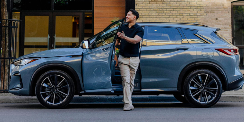 A man in a black shirt and tan pants stands next to a blue 2025 INFINITI QX50 SUV with the door open.