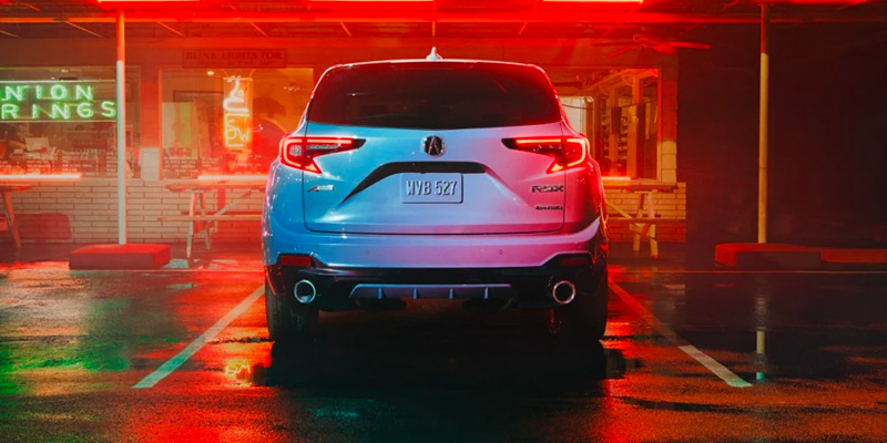 A white 2025 Acura RDX SUV parked in a neon-lit area, reflecting the colorful lights on wet pavement.