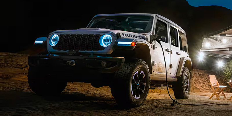 A white Jeep Rubicon parked at night with glowing headlights, near a camp setup.