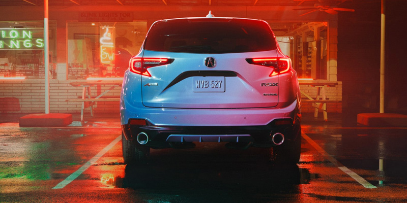 Rear view of a white Acura RDX parked at night outside a diner, illuminated by colorful neon signs. The scene reflects on the wet pavement, evoking a moody atmosphere.